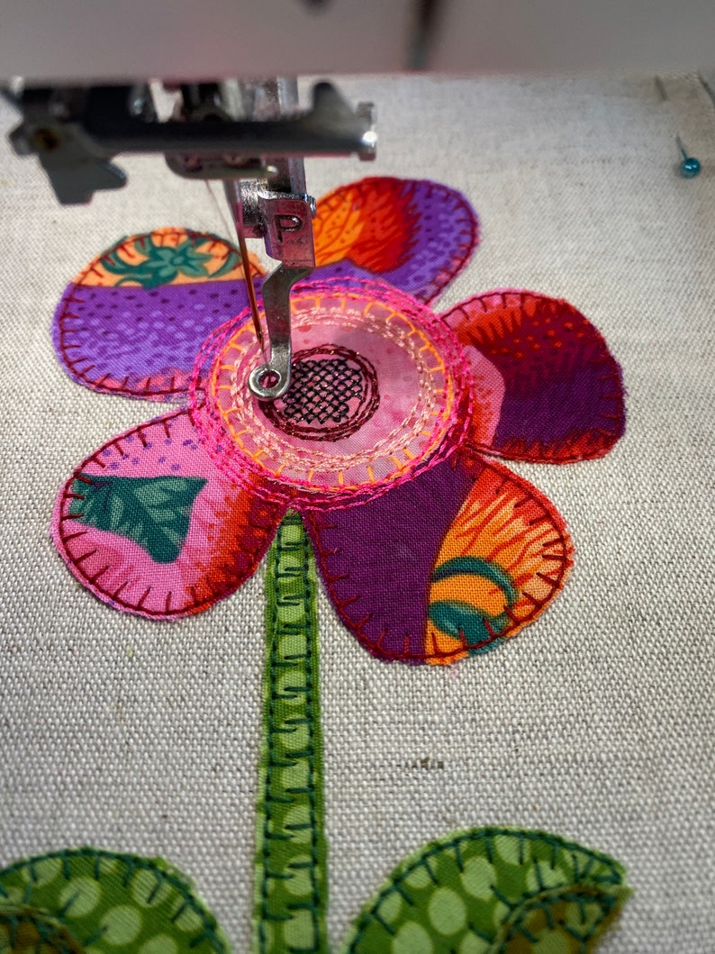 Puede incluir: Primer plano de una aguja de m&aacute;quina de coser cosiendo una flor de tela rosa y morada con hojas verdes. La flor tiene un centro rosa con un dise&ntilde;o circular.