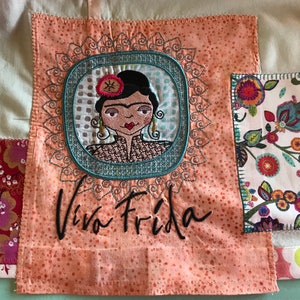 Puede incluir: Un cuadrado de tela rosa con una imagen bordada colorida de Frida Kahlo y el texto "Viva Frida".