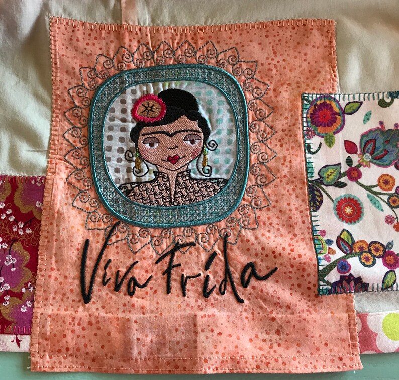 Puede incluir: Un cuadrado de tela rosa con una imagen bordada colorida de Frida Kahlo y el texto "Viva Frida".