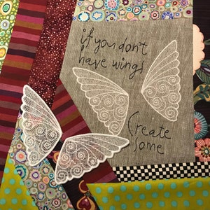 May include: A beige fabric with the text "if you don't have wings create some" embroidered in black. Two white lace wings are sewn on the fabric.
