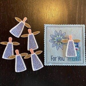 Puede incluir: Cinco peque&ntilde;os &aacute;ngeles bordados con alas doradas y vestidos con patrones azules y blancos. Los &aacute;ngeles est&aacute;n dispuestos alrededor de una tarjeta azul con una flor bordada blanca y el texto "Para Ti".