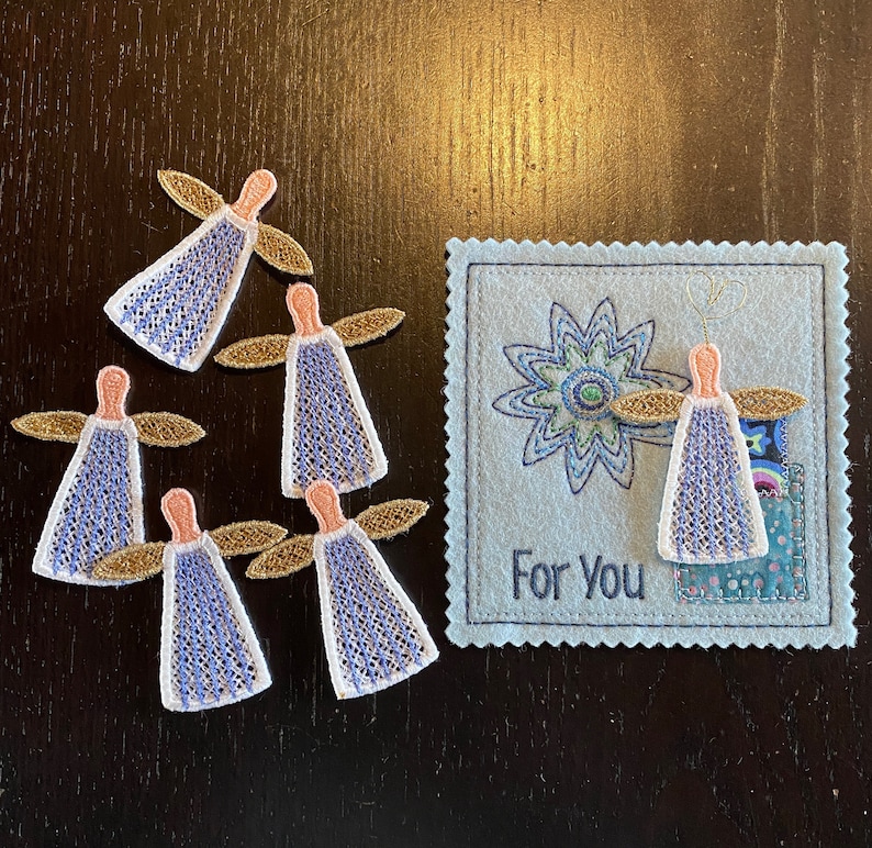 Puede incluir: Cinco peque&ntilde;os &aacute;ngeles bordados con alas doradas y vestidos con patrones azules y blancos. Los &aacute;ngeles est&aacute;n dispuestos alrededor de una tarjeta azul con una flor bordada blanca y el texto "Para Ti".