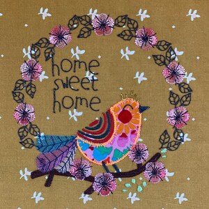 May include: Embroidered design on a brown fabric featuring a colorful bird with the text "home sweet home" surrounded by a wreath of pink flowers.