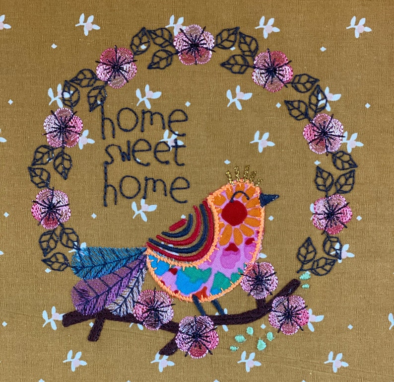 May include: Embroidered design on a brown fabric featuring a colorful bird with the text "home sweet home" surrounded by a wreath of pink flowers.