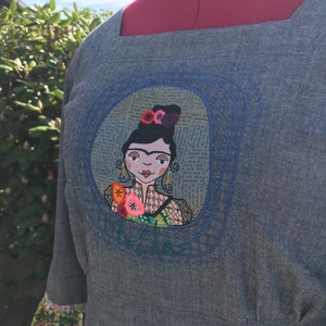 Puede incluir: Un vestido de mezclilla azul con un escote cuadrado y un dise&ntilde;o bordado de una mujer con flores en el pelo. El bordado est&aacute; rodeado por un c&iacute;rculo azul y la palabra "Frida" est&aacute; bordada debajo del c&iacute;rculo.