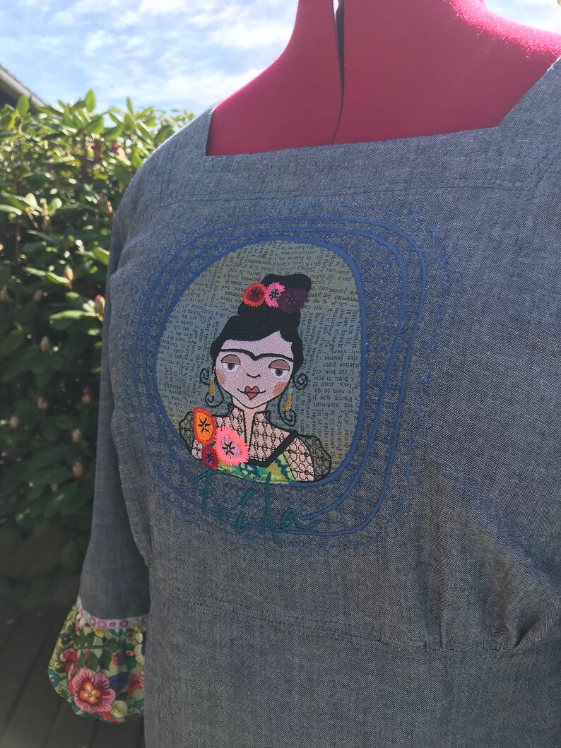 Puede incluir: Un vestido de mezclilla azul con un escote cuadrado y un dise&ntilde;o bordado de una mujer con flores en el pelo. El bordado est&aacute; rodeado por un c&iacute;rculo azul y la palabra "Frida" est&aacute; bordada debajo del c&iacute;rculo.