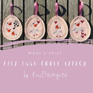 May include: Four pink felt eggs with cross stitch designs. Each egg features a white bird with different details, including hearts, musical notes, and a cross stitch pattern. The eggs are hanging from pink and white ribbon.