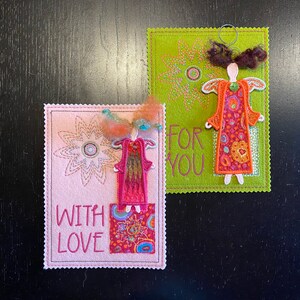 Puede incluir: Dos tarjetas de fieltro hechas a mano con &aacute;ngeles bordados y dise&ntilde;os florales. La tarjeta rosa dice "With Love" y la tarjeta verde dice "For You".