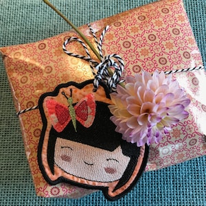 May include: A pink and white gift wrapped in gold foil paper with a pink and white floral design. The gift is tied with a black and white twine bow and has a handmade embroidered tag with a smiling girl and a butterfly.