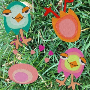 Può includere: Tre applicazioni in feltro ricamate che raffigurano un uccello verde, un uccello rosa e un fiore rosso e arancione. Le applicazioni sono circondate da erba verde.