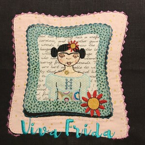 May include: Embroidered square with a Frida Kahlo design. It features a portrait of Frida, a red and yellow sun, and the words "Viva Frida" in turquoise. The piece is framed with a pink border and measures approximately 15 cm x 15 cm.