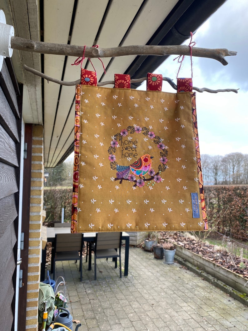 May include: A yellow fabric banner with a bird and floral wreath design. The banner has the text "home sweet home" embroidered on it. The banner is hanging from a branch.