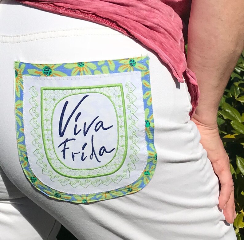 Puede incluir: Pantalones blancos con un parche bordado azul y verde en el bolsillo trasero. El parche tiene un fondo blanco con el texto "Viva Frida" en escritura cursiva azul.