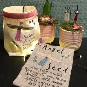Puede incluir: Una pequeña bolsa de lino beige con una cinta rosa y alas de ángel bordadas. La bolsa está iluminada desde adentro con luces de hadas y tiene el texto "This little light of mine I'm gonna let it shine" bordado en ella. Una pequeña bolsa de lino beige con el texto "Angel Seed Sow when needed Harvest all year round Fertilise with sweet, gentle words and lots of love" bordado en ella.
