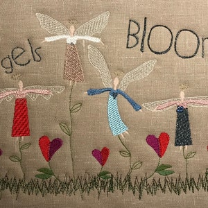 May include: Embroidered artwork featuring four angels with wings, each holding a heart. The angels are standing on stems with leaves and are surrounded by hearts. The text "Angels Bloom" is embroidered at the top of the artwork.