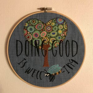 Puede incluir: Arte de bordado en aro con fondo azul, un árbol en forma de corazón con círculos coloridos, un tronco marrón y el texto "Hacer el bien vale la pena intentarlo". Una abeja azul y amarilla con ojos blancos vuela debajo del árbol.