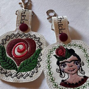 May include: Two white leather keychains with embroidered designs. One keychain features a red rose with green leaves and the other features a portrait of Frida Kahlo with a red rose in her hair. Both keychains have the name "Frida" embroidered on the top.