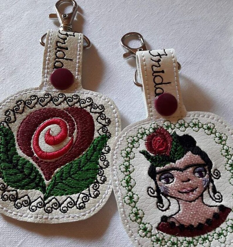 May include: Two white leather keychains with embroidered designs. One keychain features a red rose with green leaves and the other features a portrait of Frida Kahlo with a red rose in her hair. Both keychains have the name "Frida" embroidered on the top.