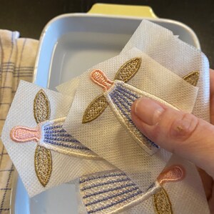 Puede incluir: Tres aplicaciones de &aacute;ngel de tela blanca con bordados dorados y rosados. Las aplicaciones est&aacute;n en un recipiente de pl&aacute;stico transparente lleno de agua.