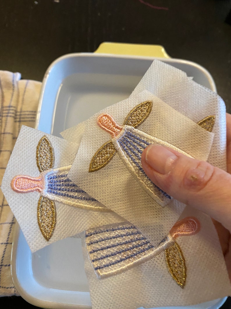 Puede incluir: Tres aplicaciones de &aacute;ngel de tela blanca con bordados dorados y rosados. Las aplicaciones est&aacute;n en un recipiente de pl&aacute;stico transparente lleno de agua.