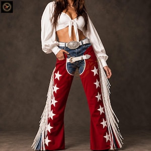 May include: A white long-sleeved cropped blouse with a front tie, worn with blue denim jeans and red chaps featuring white star patterns and long white fringe. A white leather belt with a large silver buckle is worn over the jeans.