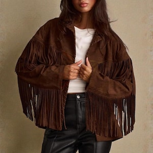 Puede incluir: Chaqueta de ante marrón con flecos largos en las mangas y el pecho. La chaqueta se lleva sobre una camiseta blanca y se combina con pantalones de cuero negros. La persona lleva gafas de sol negras.