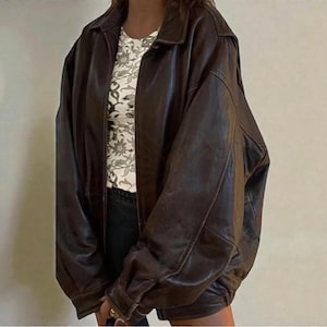 May include: A dark brown leather jacket with a collared neckline and a zip-up front. The jacket has a relaxed fit and features a shirt underneath with a floral pattern. The jacket is a classic outerwear piece.
