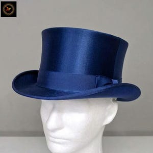 Puede incluir: Un sombrero de copa azul marino profundo con un acabado suave y satinado. El sombrero presenta una copa cilíndrica alta y un ala curva. Una cinta azul marino a juego rodea la base de la copa, añadiendo un detalle sutil al diseño general.