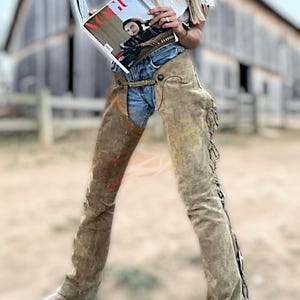 Op de afbeelding: Tan suède chaps met franjes, gedragen over een blauwe spijkerbroek. De chaps hebben een klassiek western design. De persoon houdt een tijdschrift vast met het woord "VOGUE" op de cover. De achtergrond toont een rustieke houten schuur.
