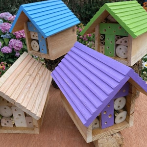 Puede incluir: Cuatro casas de insectos de madera con tejados coloridos. Una tiene un tejado azul, otra verde, una tercera morada y la última es de madera natural. Cada casa tiene un marco de madera y está llena de materiales naturales.