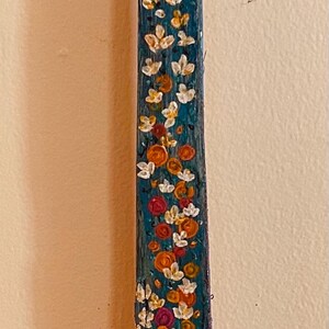 Puede incluir: Una pieza de arte de madera pintada a mano con un fondo turquesa y acentos florales coloridos. La parte superior tiene un diseño marrón oscuro texturizado, y la parte inferior presenta un pomo decorativo con dibujos. La pieza mide unos 76 cm de alto.