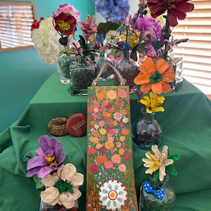 May include: A vibrant display of handmade floral art and decor. Features a wooden floral painting, decorative stones with text, and various flowers in glass bottles. The color palette includes greens, oranges, pinks, and blues.