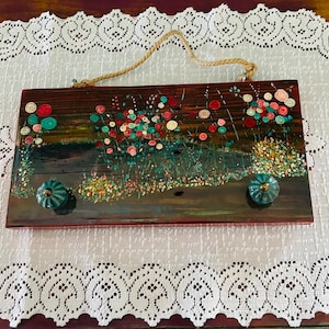 May include: A rectangular wooden wall hanging with a painted floral design in red, teal, and yellow. The artwork features two teal knobs and a natural rope for hanging. The background is a dark brown.
