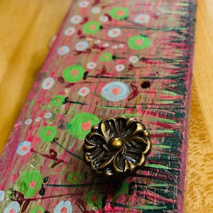 May include: A rectangular wooden decorative piece, painted with pink, green, and white floral designs. It features two ornate bronze-colored knobs. The artwork has a rustic, handmade aesthetic.