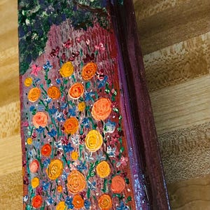 May include: Hand-painted wooden wall art featuring a vibrant floral scene with orange, yellow, and blue flowers. The artwork has a dark green and purple background with a decorative knob at the bottom. A natural rope is attached for hanging.