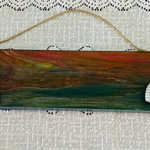 May include: A rectangular wooden wall hanging with a painted sunset design in shades of red, orange, and green. Two white seashell accents are attached. A jute rope is attached for hanging.
