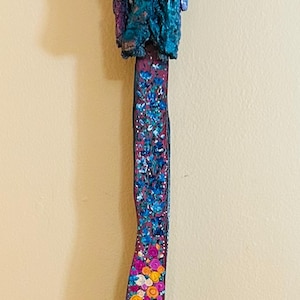 May include: A tall, vertical sculpture with a textured, turquoise top section. The stem is painted with colorful floral patterns, transitioning to a base with a decorative, patterned sphere. The artwork is mounted on a neutral wall.