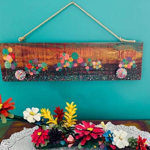 May include: A rectangular wooden wall art piece with a colorful abstract floral design. The artwork features a textured background with various colorful circular shapes and a rope for hanging. Below, a floral arrangement with red, white, and yellow flowers.
