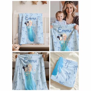 Puede incluir: Manta de beb&eacute; azul claro con Mickey Mouse y Elsa de Frozen, con el nombre "Emma". La manta tiene un patr&oacute;n de lazos y se muestra en m&uacute;ltiples entornos, incluyendo una cuna y sostenida por una persona.