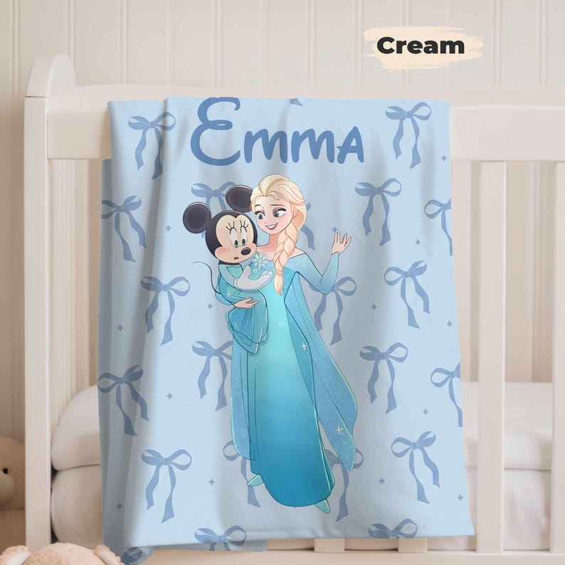 Puede incluir: Una manta de beb&eacute; azul claro con el nombre "Emma" impreso. La manta presenta un patr&oacute;n de lazos azul claro y una ilustraci&oacute;n de dibujos animados de Elsa de Frozen sosteniendo a Mickey Mouse. Se muestra en una cuna blanca.