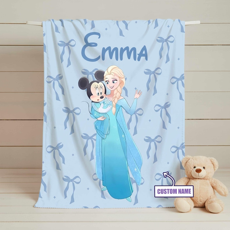 Puede incluir: Manta de beb&eacute; azul claro con una ilustraci&oacute;n de dibujos animados de Elsa de Frozen sosteniendo a Mickey Mouse. La manta tiene un patr&oacute;n de lazos y el nombre "Emma" impreso. Un osito de peluche est&aacute; al lado de la manta.