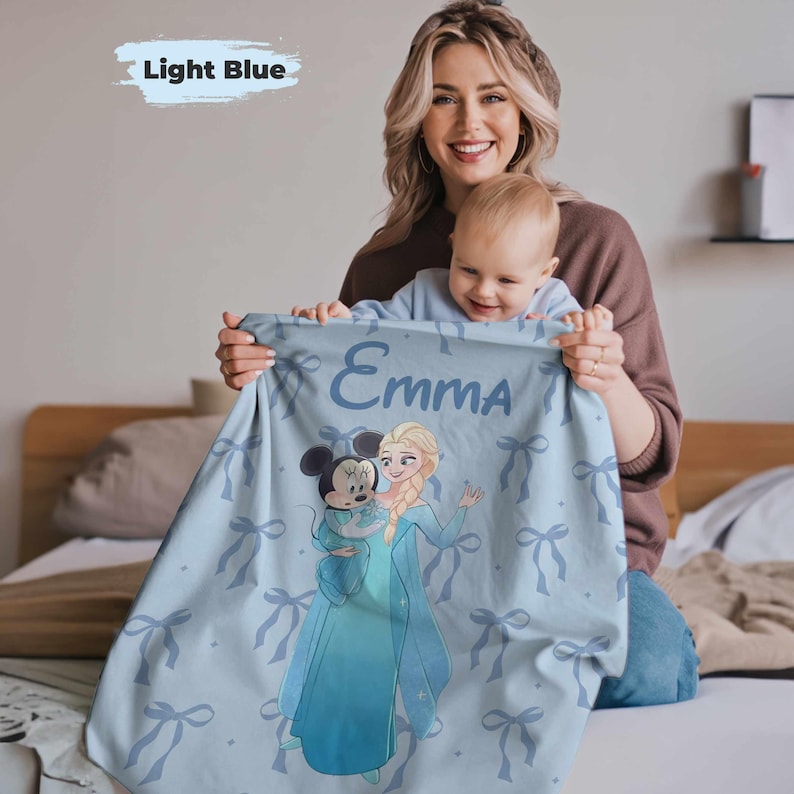 Puede incluir: Manta de beb&eacute; azul claro con una ilustraci&oacute;n de dibujos animados de Elsa y Minnie Mouse. La manta tiene un patr&oacute;n de lazo repetido y el nombre "Emma" impreso en azul claro.