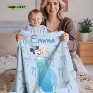 Puede incluir: Una manta de beb&eacute; azul claro con el nombre "Emma" impreso. La manta tiene un patr&oacute;n de lazos y un gr&aacute;fico de Mickey Mouse y Elsa de Frozen. La manta est&aacute; sostenida por una mujer y un beb&eacute;.