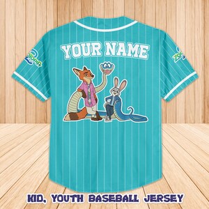 Op de afbeelding: Turquoise honkbalshirt met witte krijtstrepen, met stripfiguren en de tekst "YOUR NAME" op de rug. De trui heeft witte biezen op de kraag en mouwen, en de tekst "KID, YOUTH BASEBALL JERSEY" staat onderaan.