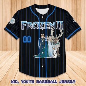 Puede incluir: Camiseta de b&eacute;isbol negra con finas rayas y detalles azules. La camiseta presenta el texto "FROZEN II" y un gr&aacute;fico de personajes de la pel&iacute;cula. El n&uacute;mero "00" est&aacute; en la parte delantera. El texto "KID, YOUTH BASEBALL JERSEY" est&aacute; en la parte inferior.