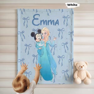 Puede incluir: Una manta azul claro con una ilustraci&oacute;n de dibujos animados de Elsa y Minnie Mouse, con el nombre "Emma". La manta tiene un patr&oacute;n de lazos azul claro. Un osito de peluche est&aacute; a la derecha.