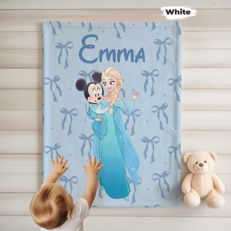 Puede incluir: Una manta azul claro con una ilustraci&oacute;n de dibujos animados de Elsa y Minnie Mouse, con el nombre "Emma". La manta tiene un patr&oacute;n de lazos azul claro. Un osito de peluche est&aacute; a la derecha.