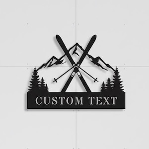 Może przedstawiać: Czarna metalowa grafika ścienna przedstawiająca skrzyżowane narty, góry i sosny. Projekt zawiera tekst "CUSTOM TEXT" na prostokątnej podstawie. Grafika jest zamontowana na białej ścianie z ośnieżonym górskim tłem.