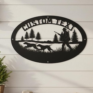 Könnte beinhalten: Schwarze Metall-Wandkunst mit einer Jagdszene mit Hunden, Bäumen und einem Jäger mit Gewehr. Das ovale Design enthält den Text "CUSTOM TEXT". Die Kunst wird an einer weißen Wand mit Lamellen ausgestellt, mit kleinen Topfbäumen auf Regalen.