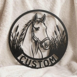 Könnte beinhalten: Ein rundes Wandbild aus schwarzem Metall mit einem Scherenschnitt-Design eines Pferdekopfes mit Zaumzeug, umgeben von hohem Gras und Schilfkolben. Das Wort CUSTOM ist in Großbuchstaben am unteren Rand des kreisförmigen Rahmens eingeprägt.